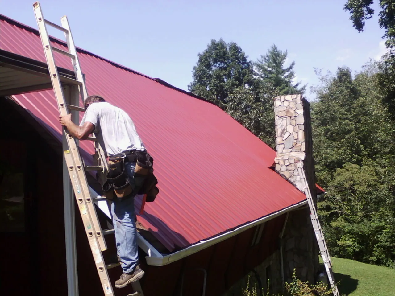 Roofer on a ladder beside a red metal roof and stone chimney — Soffit & Fascia Repair by Roofing Brothers in Buchtel